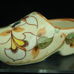 May include: A white ceramic Dutch shoe with a floral design in shades of brown, yellow, and green. The shoe is a small decorative piece with a rounded shape and a small opening at the top.