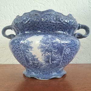 Może przedstawiać: Niebiesko-biała ceramiczna miska z dwoma uchwytami, przedstawiająca szczegółowy krajobraz z drzewami i listowiem z przodu, otoczona geometrycznym wzorem oraz falistym brzegiem z wypukłymi detalami kwiatowymi.