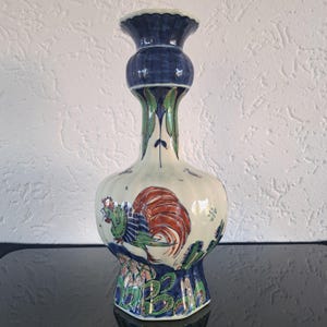 May include: A white ceramic vase with a blue and green floral design and a rooster painted on the side. The vase has a wide base and a narrow neck.
