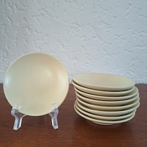 May include: A set of small, round, pale yellow ceramic plates. One plate is displayed upright on a clear plastic stand, while a stack of seven identical plates is positioned to the right, showcasing their smooth, matte finish and simple design.