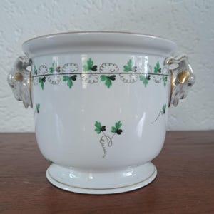 May include: A white porcelain planter featuring a green and black leaf pattern around the rim and scattered on the body. The vessel includes two sculpted animal head handles on the sides, accented with gold trim on the ears and base rim.