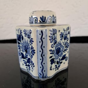 May include: A white ceramic jar with a blue and white floral design. The jar has a lid and is decorated with a traditional Dutch Delftware pattern.