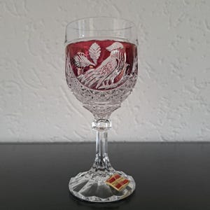 Puede incluir: Una copa de vino de cristal transparente con interior rojo rubí. La copa presenta un diseño grabado de un pájaro y follaje. El tallo y la base también son de cristal, y la base tiene una pequeña pegatina. La copa está sobre una superficie oscura.