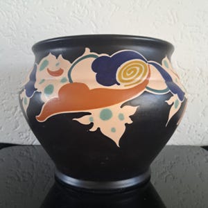 Könnte beinhalten: Eine schwarze Keramikvase mit breiter Öffnung und rundem Körper. Die Vase ist mit einem farbenfrohen Blumenmuster in Orange, Blau und Creme verziert. Das Design umfasst stilisierte Blätter und wolkenartige Formen.