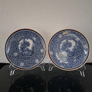 Puede incluir: Dos platos de cerámica azul y blanco con un borde marrón y un diseño de árbol en el centro. Los platos están sobre soportes blancos.