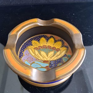 Può includere: Un posacenere rotondo in metallo con finitura color bronzo e accenti arancioni lungo il bordo. Il centro presenta un inserto in smalto colorato che raffigura un fiore giallo stilizzato con dettagli blu e viola su uno sfondo blu scuro puntinato.