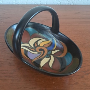 May include: An oval ceramic basket with a black exterior and a high arched handle. The interior features a hand-painted floral motif in shades of cream, orange, blue, and purple against a black background, creating a bold contrast.