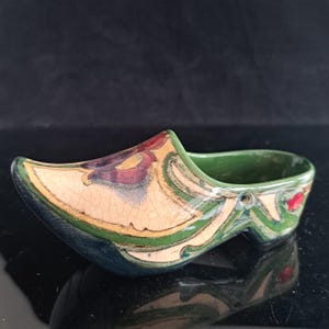 Op de afbeelding: Een decoratief object van keramiek in de vorm van een klomp met een crèmekleurige basis en een craquelé glazuur. De buitenkant is versierd met wervelende patronen in donkergroen, goud en rood, terwijl de binnenkant effen donkergroen is.
