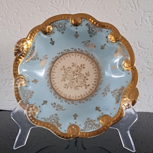 May include: A vintage, light blue decorative dish with a scalloped gold rim. The center features a cream-colored circle with a gold floral design. Gold filigree and floral accents adorn the blue surface. The dish is displayed on a clear plastic stand.