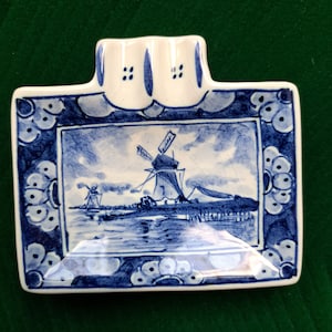 May include: A rectangular, blue and white ceramic ashtray featuring a windmill scene. The ashtray has a raised section with two slots and is decorated with a floral border. The windmill scene depicts a body of water and a cloudy sky.