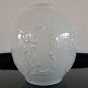 Può includere: Vaso in ceramica bianca con un intricato design floreale in rilievo. Il vaso ha una forma rotonda e una finitura liscia e lucida.