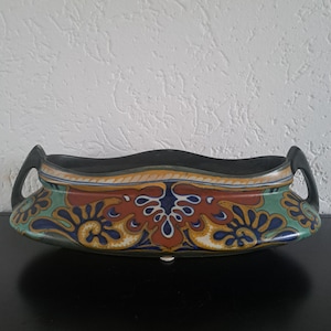 May include: A ceramic planter with a black rim and handles, featuring a colorful floral design in shades of red, blue, yellow, and green. The planter has a curved shape and sits on small feet.