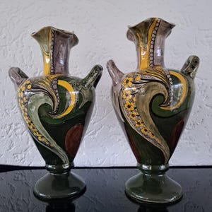 Peut inclure: Deux vases en céramique de couleur verte, jaune et marron avec des motifs floraux et géométriques complexes. Les vases ont une base large et un col étroit.
