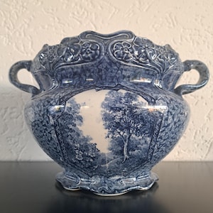 Könnte beinhalten: Ein blau-weißer Keramik-Pflanztopf mit einer detaillierten Landschaftsszene. Der Pflanztopf hat zwei Griffe und einen gewellten Rand mit floralen Details. Die Außenseite weist ein strukturiertes Muster auf, und die Innenseite ist weiß.