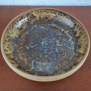 May include: A round ceramic bowl featuring a reactive glaze with a dark brown and black centre, transitioning into a lighter tan and beige rim. The interior surface displays a mottled, textured appearance with subtle blue and white highlights.