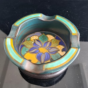 Può includere: Un posacenere rotondo in ceramica con un bordo verde acqua scuro caratterizzato da accenti e tacche gialle. La base interna presenta un vivace motivo floreale con un grande fiore blu, foglie verdi e dettagli circolari gialli.