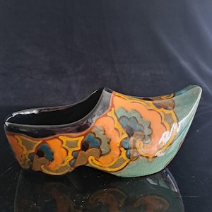 Puede incluir: Un objeto decorativo de cerámica con forma de zueco que presenta un acabado brillante con un patrón vibrante de remolinos florales en naranja, verde azulado y amarillo. El interior es negro sólido y la superficie exterior muestra una textura craquelada.