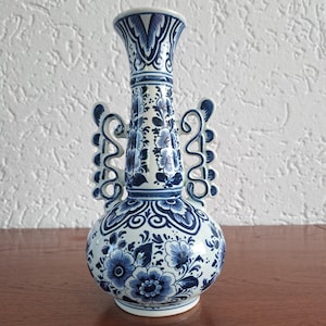 Può includere: Un vaso in ceramica blu e bianco, in stile Delft, con base arrotondata e collo sottile. Il vaso è decorato con motivi floreali e volute. Ha due manici decorativi ed è alto circa 25 cm.