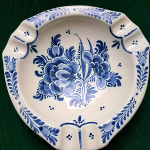 May include: A white ceramic ashtray with a blue and white floral design. The ashtray has a rounded shape with three indentations for cigarettes.