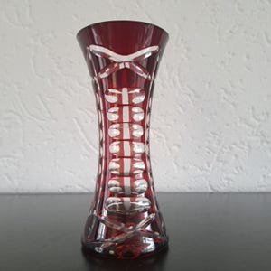 May include: A ruby red glass vase with a flared top and a patterned design. The vase features a series of vertical, oval-shaped cutouts, and crisscross patterns. The vase is a decorative item.