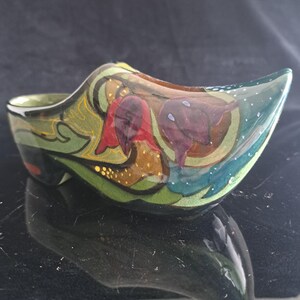 Puede incluir: Un zueco decorativo de cerámica con acabado brillante y motivos florales pintados a mano en tonos rojo intenso, morado y verde. El adorno en forma de zapato presenta patrones intrincados, detalles en tono dorado y una punta afilada.