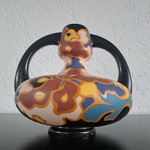 Könnte beinhalten: Eine Keramikvase mit schwarzem Sockel und Griffen. Die Vase hat einen cremefarbenen Körper mit einem Blumenmuster in Braun, Gelb und Blau. Die Griffe sind schwarz und die Vase hat eine einzigartige, abgerundete Form.