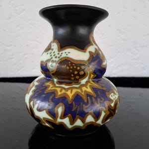 May include: A small, black ceramic vase with a bulbous body and a narrow neck. The vase is decorated with a colorful, abstract design in shades of blue, green, yellow, and brown.