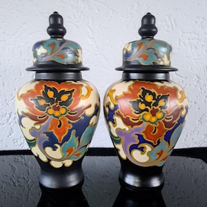 Può includere: Un paio di vasi in ceramica nera con coperchi, decorati con motivi floreali colorati in tonalità di rosso, arancio, giallo, blu, verde e viola. I vasi hanno un design cinese tradizionale.