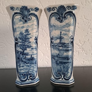 Puede incluir: Dos jarrones de cerámica azul y blanca a juego. Cada jarrón presenta un paisaje, uno con un árbol y un barco, el otro con un molino de viento y un barco. Los jarrones tienen forma acampanada y adornos azules decorativos.