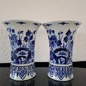 Può includere: Due vasi in ceramica bianca con motivi floreali blu e bianchi. I vasi hanno una parte superiore svasata e una base rotonda. I motivi floreali sono intricati e dettagliati.