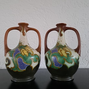 May include: Two antique ceramic vases with brown handles and rims. The vases have a cream-colored body with a floral design in yellow, green, and blue. The base of the vases is dark green.