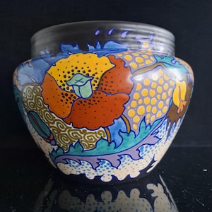 May include: A rounded ceramic vase featuring a dark black rim and a vibrant, multi-coloured floral pattern. The design includes large orange and yellow poppy-like flowers, green leaves, and intricate blue and gold dotted and swirling textures.