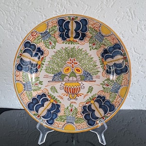 Könnte beinhalten: Dekorative Keramikplatte mit handgemaltem Blumendesign. Die Platte zeigt eine zentrale Anordnung von Früchten und Blättern, umgeben von blauen und gelben Blumenmotiven. Der Rand ist mit einem gelben Rand versehen.