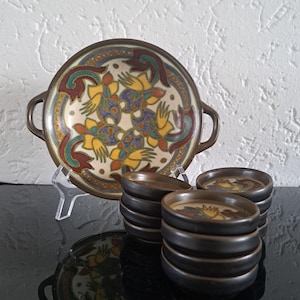 Puede incluir: Una fuente de cerámica con asas y un juego de posavasos a juego. La fuente y los posavasos presentan un fondo color crema con un diseño floral en amarillo, azul y granate. Los bordes son de color marrón oscuro.