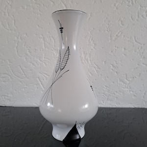 Puede incluir: Un jarrón de cerámica blanco con borde y base negros. El jarrón presenta delicados dibujos lineales negros de espigas de trigo y formas abstractas. El jarrón tiene un cuerpo redondeado y una parte superior acampanada, de unos 25 cm de altura.