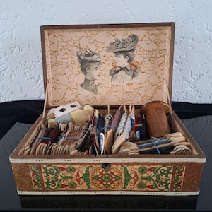 Op de afbeelding: Een antieke houten naaidoos met een deksel met een bloemenpatroon. De doos is gevuld met spoelen garen, knopen en andere naaigerei.