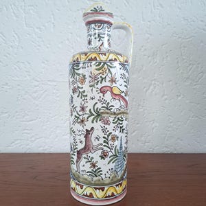 May include: A cylindrical ceramic bottle with a handle and a matching stopper, featuring a hand-painted design of a brown deer, a pink bird, and various floral motifs in shades of yellow, green, and red against a white background.