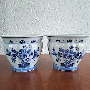 May include: Two white ceramic planters featuring a blue floral pattern with intricate petals and leaves. Each planter has a fluted, rounded body with a wide rim and a solid blue base, showcasing a classic hand-painted style design.