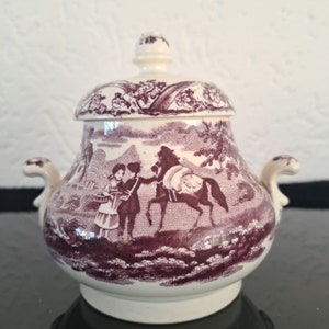 May include: A white ceramic sugar bowl with a lid, decorated with a purple floral pattern and a scene of two people and a horse. The bowl has two handles on the sides.