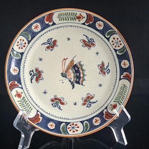 May include: A round ceramic plate with a cream-colored base featuring a central butterfly illustration. The plate has a blue border decorated with red and green floral motifs and small star-like accents. The surface has a visible crackle glaze finish.