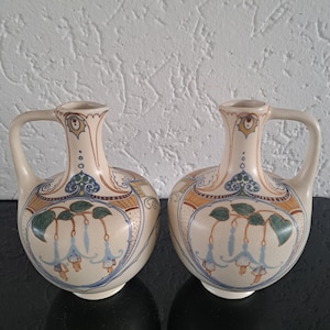 May include: Two cream-colored ceramic vases with handles. Each vase features a floral design with blue, green, and orange accents. The vases have a bulbous body and a narrow neck, with a handle on each side.