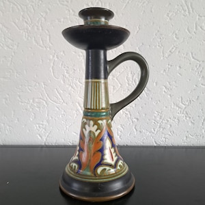 Può includere: Un portacandele in ceramica con base e parte superiore marrone scuro, stelo nero e manico. La base presenta un colorato motivo floreale con accenti arancioni, blu e verdi. Il portacandele è alto circa 20 cm.