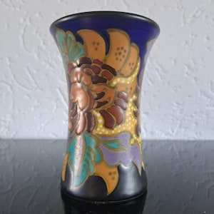 Puede incluir: Un pequeño jarrón decorativo con borde y base azul oscuro. El jarrón presenta un diseño floral en tonos marrón, naranja, amarillo, verde azulado y morado. El diseño está sobre un fondo negro.