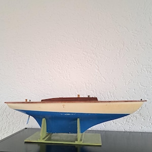 May include: A wooden model sailboat with a cream-colored hull, blue bottom, and brown deck. The model is mounted on a green base and sits on a black surface. The sailboat has a long, sleek design.