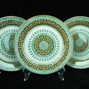Puede incluir: Tres platos decorativos con fondo blanco y diseño geométrico verde y dorado. Los platos se exhiben sobre soportes transparentes sobre un fondo negro. Los platos tienen un patrón de greca.