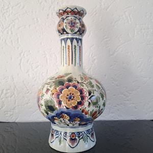 Puede incluir: Un jarrón de cerámica blanca con un cuerpo bulboso y un cuello alto y delgado. El jarrón está decorado con un patrón floral colorido en tonos de azul, amarillo, rosa y verde.