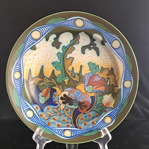 Puede incluir: Un plato de cerámica redondo con un motivo floral central en tonos azul, naranja y verde, rodeado por un borde de puntos amarillos y óvalos a rayas azules. El fondo incluye un patrón amarillo punteado y formas de hojas de color verde oscuro.