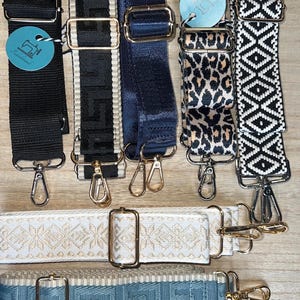 Könnte beinhalten: Auswahl an verstellbaren Taschenriemen in verschiedenen Designs. Enthält Schwarz, Marineblau, Leopardenmuster und geometrische Muster. Die Riemen verfügen über Metallbeschläge und sind für Handtaschen konzipiert. Einige Riemen haben den Text "Tadahhandmade" auf einem Anhänger.