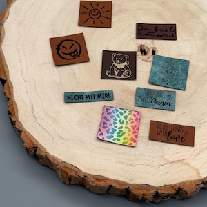 Könnte beinhalten: Eine Sammlung rechteckiger Lederaufnäher mit verschiedenen Designs und Texten, darunter eine lächelnde Sonne, ein Bär und der Satz „DO ALL THINGS WITH love“. Die Aufnäher sind auf einer Holzoberfläche angeordnet.