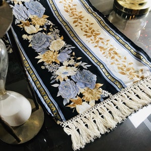 May include: A black and white table runner with a floral pattern in shades of blue, yellow, and brown. The runner has a white fringe trim.